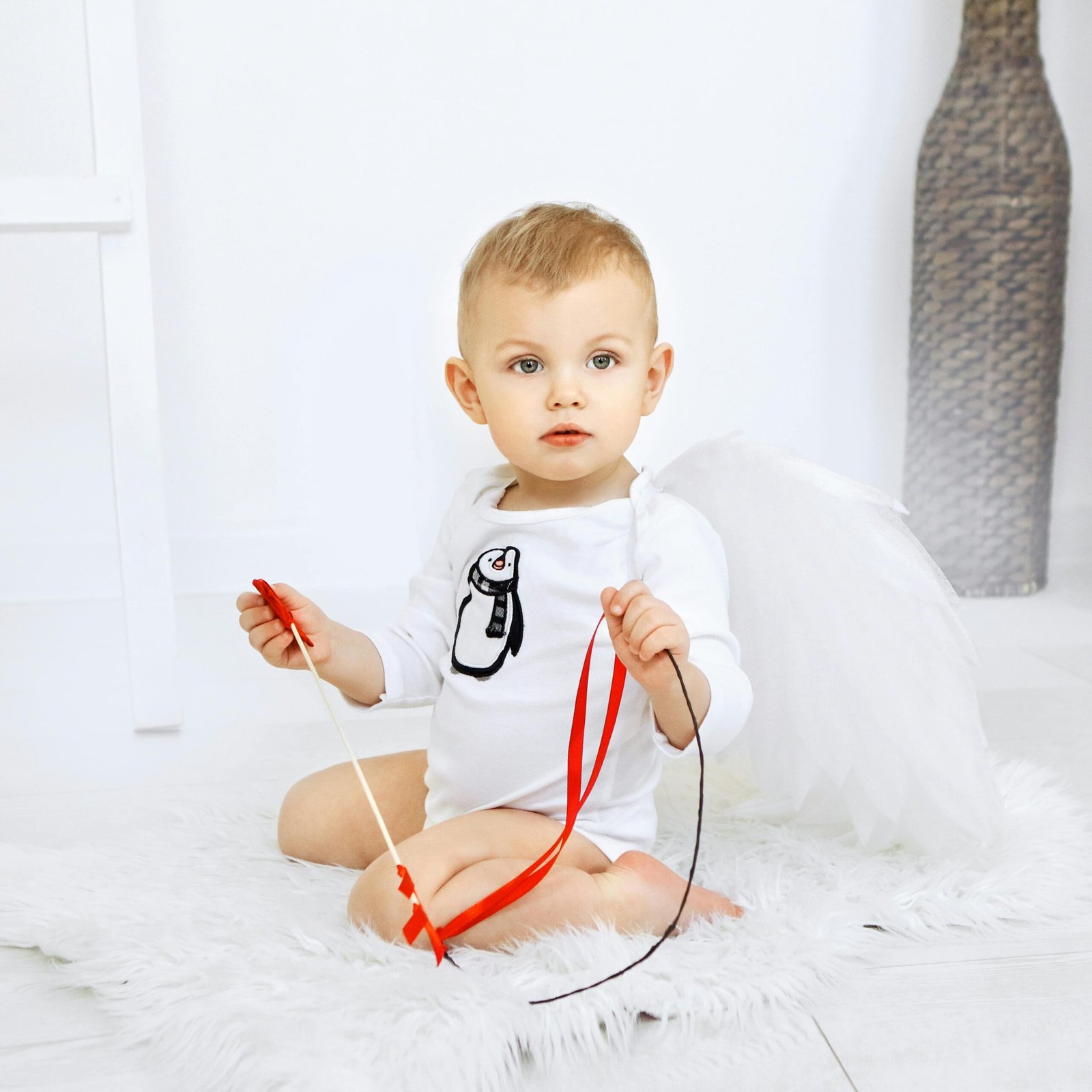 Cute baby sitting on a rug with ribbons, wearing angel wings, indoors.